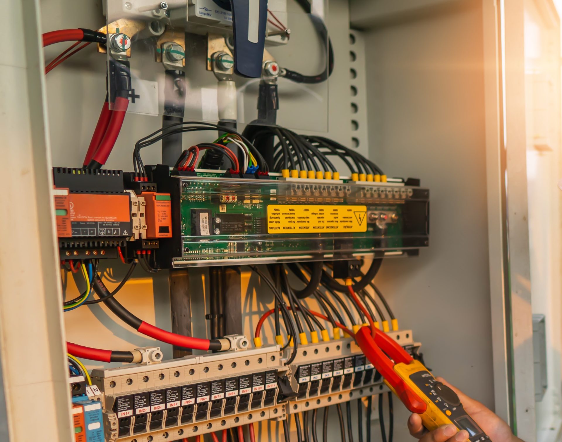 An open electrical cabinet with an isolator switch, a row of breakers and terminals, and a hand holding a clamp meter around one of the cables