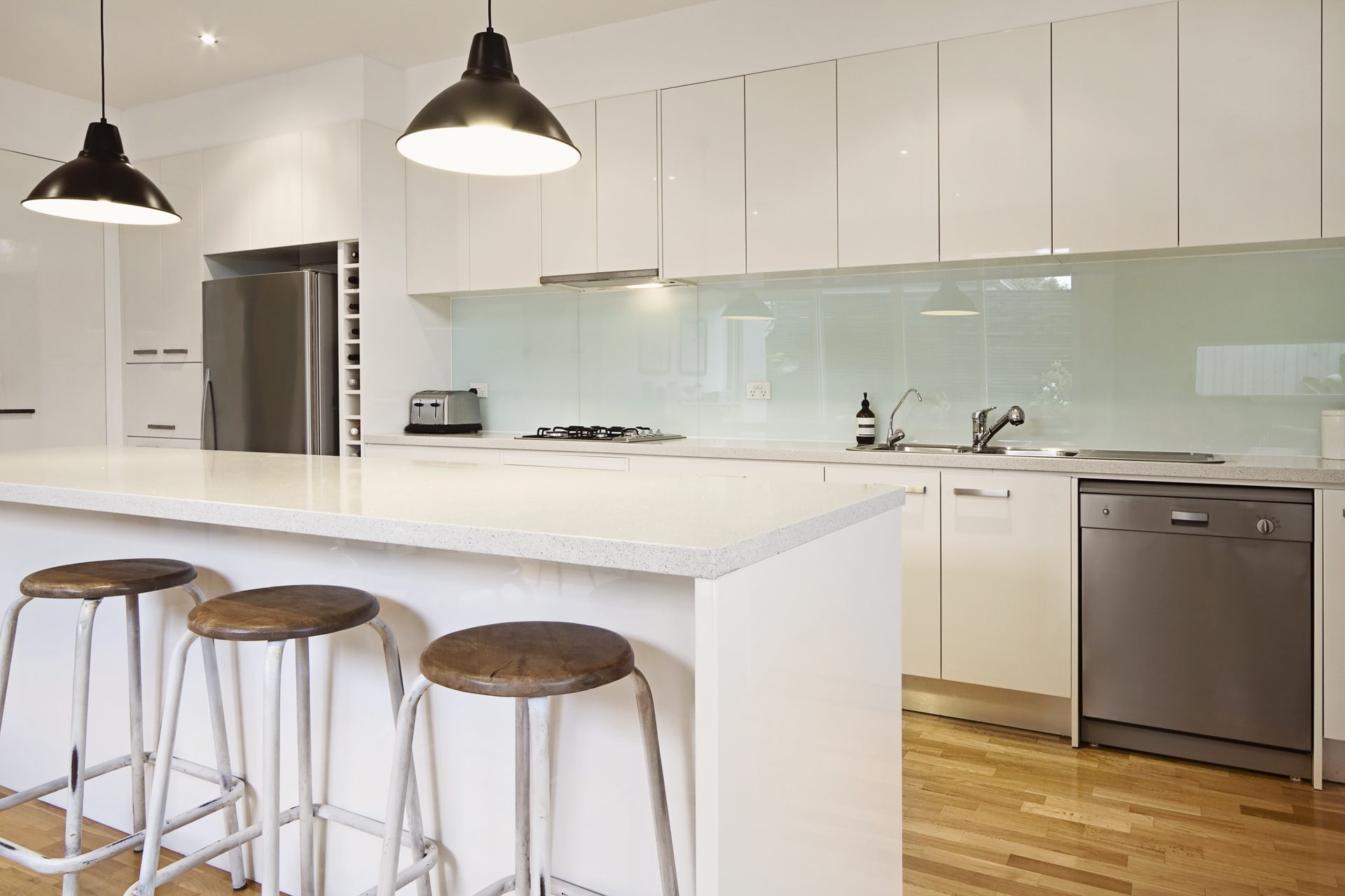 A white kitchen with two black pendant lights over an island bench, three timber-topped stools and a timber floor